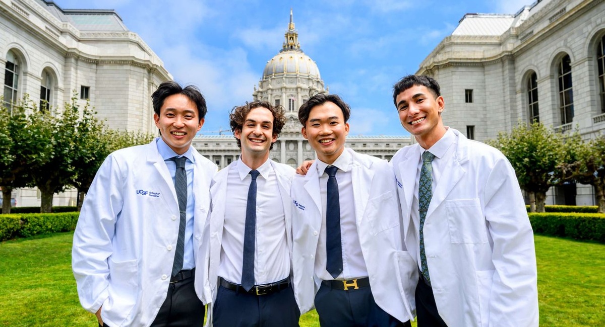 Four smiling UCSF School of Medicine students wearing white coats pose together in front of San Francisco City Hall.