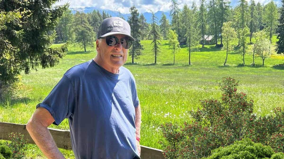 Man standing outdoors in a grassy field with trees and mountains in the background