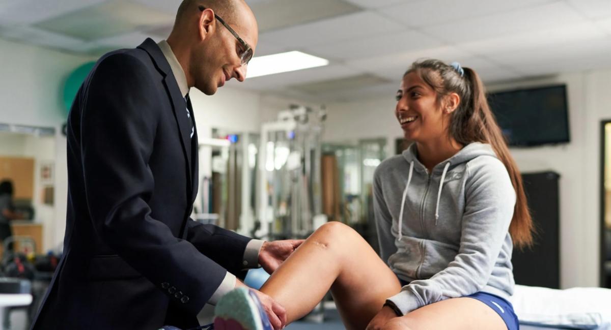 Health care provider examines a young athlete’s knee during a rehabilitation appointment as she smiles and talks with him.
