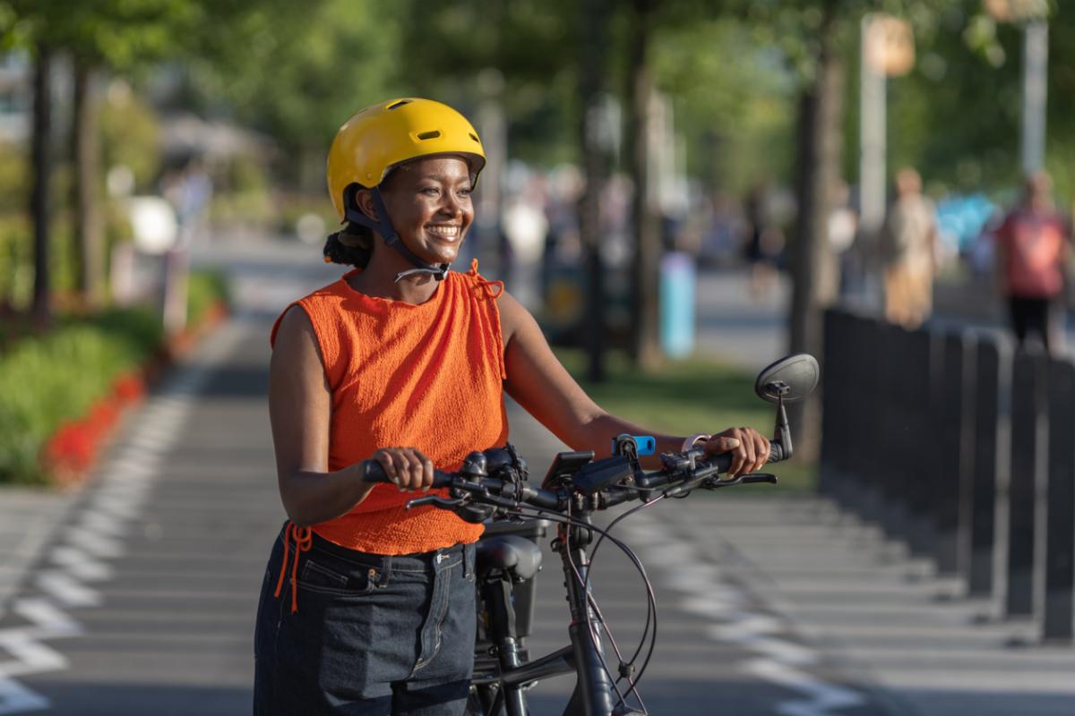 Person wearing a yellow helmet stands with an e-bike on a tree-lined bike path, smiling.