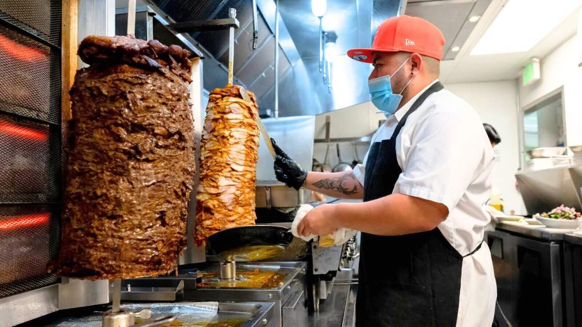 Man in kitchen cuts meat from a large blazing skewer.
