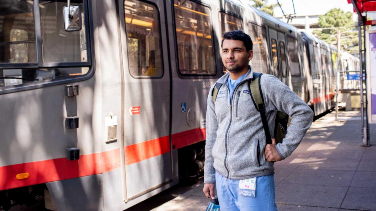 A medical student stands in front of a San Francisco MUNI rail train.