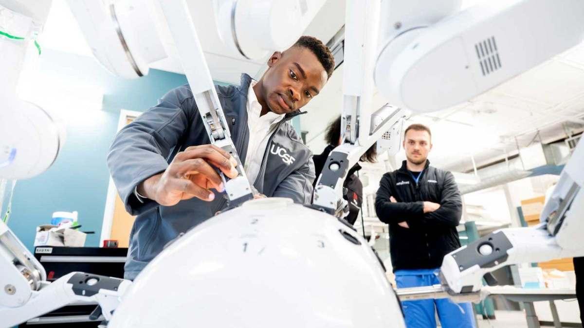 Young man in lab coat looks intensely at large white robotic machine in a bright room.