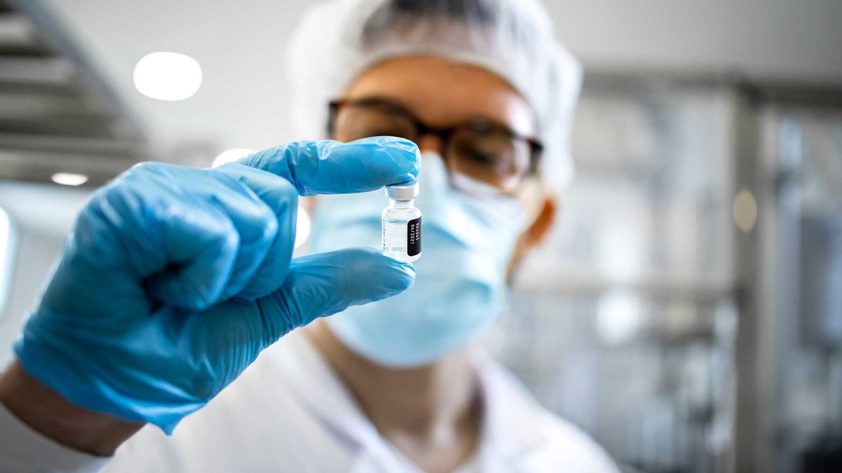Scientist wearing protective gloves, a mask, and a hair covering examines a small vial in a laboratory.