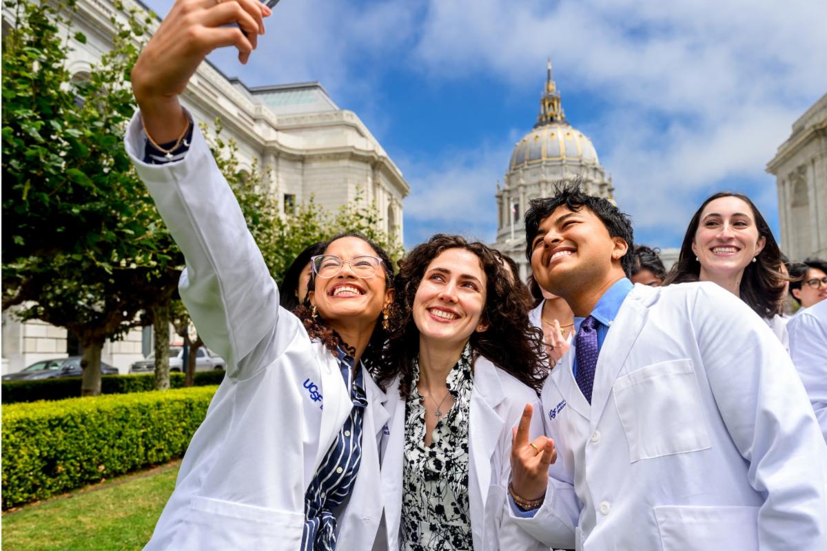 UCSF medical students wearing white coats smile together while taking a selfie in front of San Francisco City Hall.