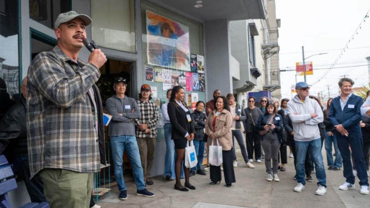 Man on microphone stands in front of theater talking to a group holding canvas bags.