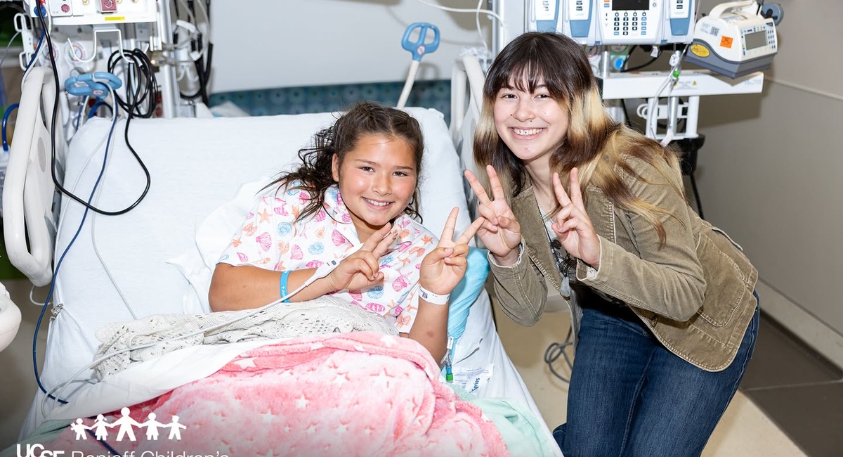 Smiling child in a hospital bed and Olympic figure skating champion — and Oakland native — Alysa Liu pose with peace signs in a UCSF Benioff Children’s Hospitals patient room.