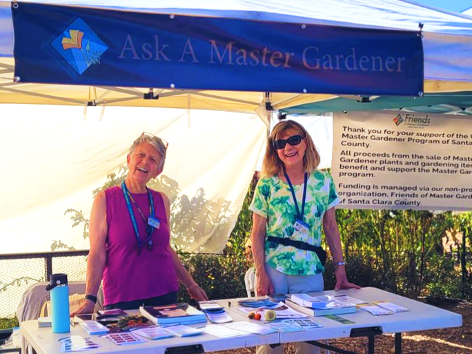 Two women who are Master Gardeners standing behind a table with a blue banner above them which reads “Ask a Master Gardener”