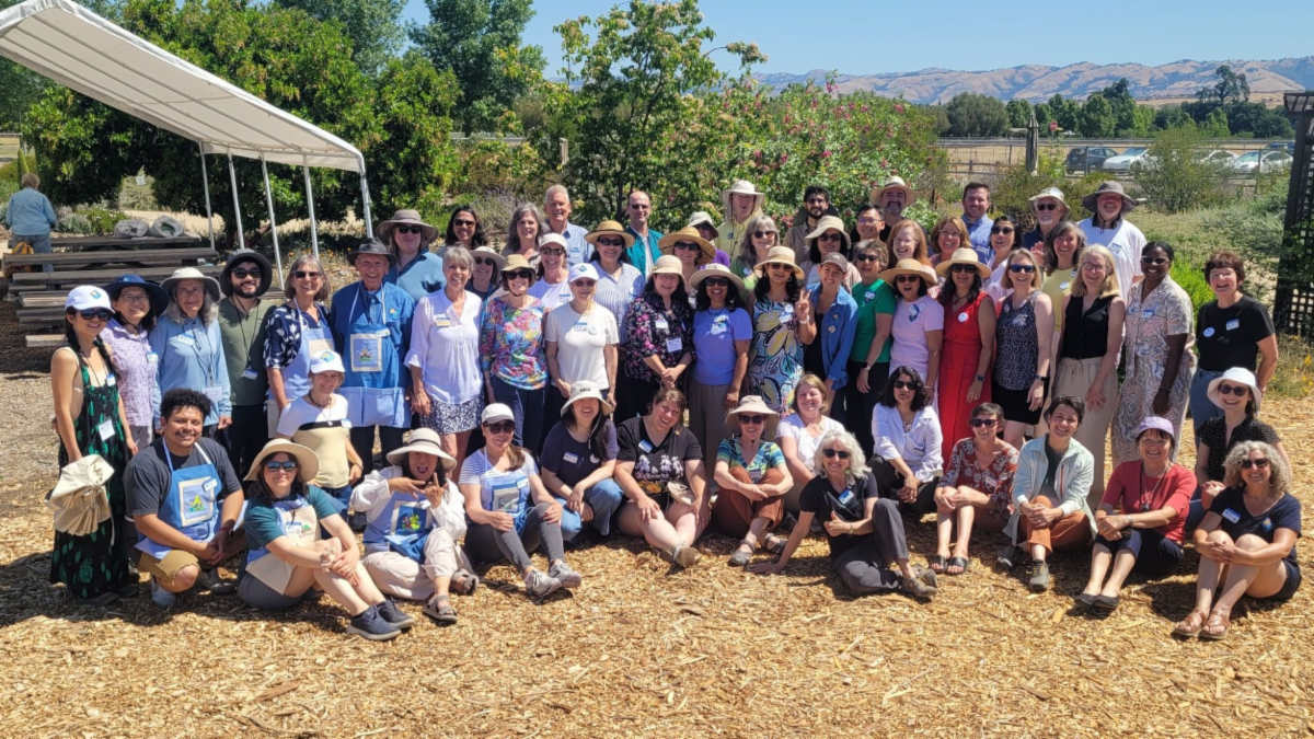 A diverse group of about 50 people smiling and posing outdoors in a park, wearing hats and gathered in front of lush greenery and a clear sky.
