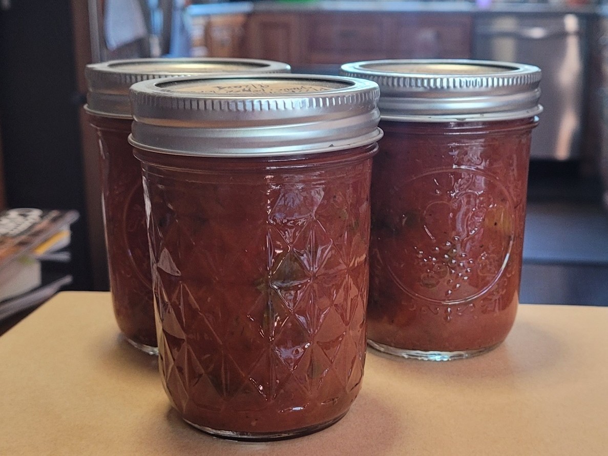 Three glass jars of reddish-brown chunky pizza sauce preserved with silver lids on a kitchen counter.