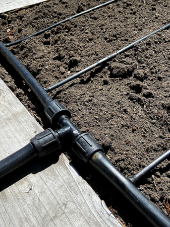 Photograph showing a close-up of a drip irrigation system installed in a garden bed with dark, loose soil. Black plastic tubing with connectors runs parallel along soil surface, designed to deliver water efficiently to plants.