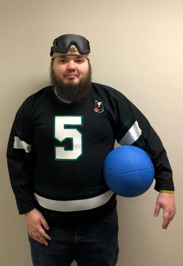 Photo of Jordan Weeden standing with a goalball, wearing a goalball jersey and blackout goggles on his forehead