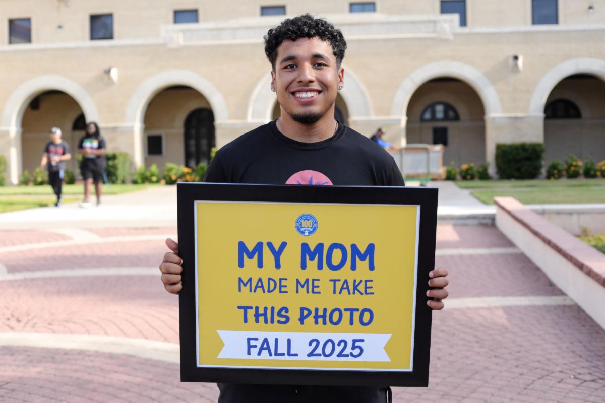 A student smiles holding a sign that says, "My Mom Made Me Take This Photo Fall 2025"