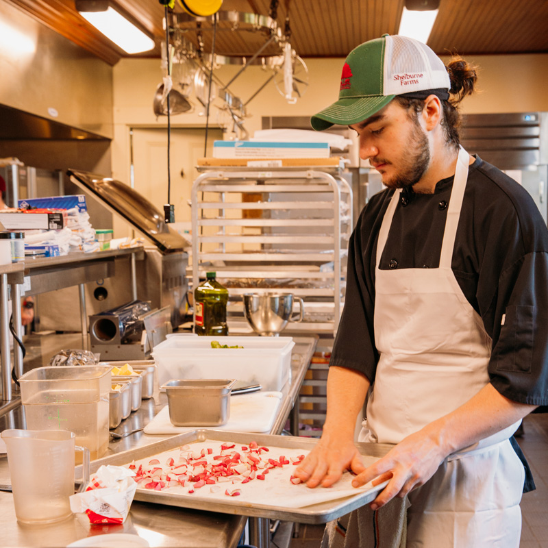 A person in a professional kitchen wearing a white apron is preparing food on a metal table.