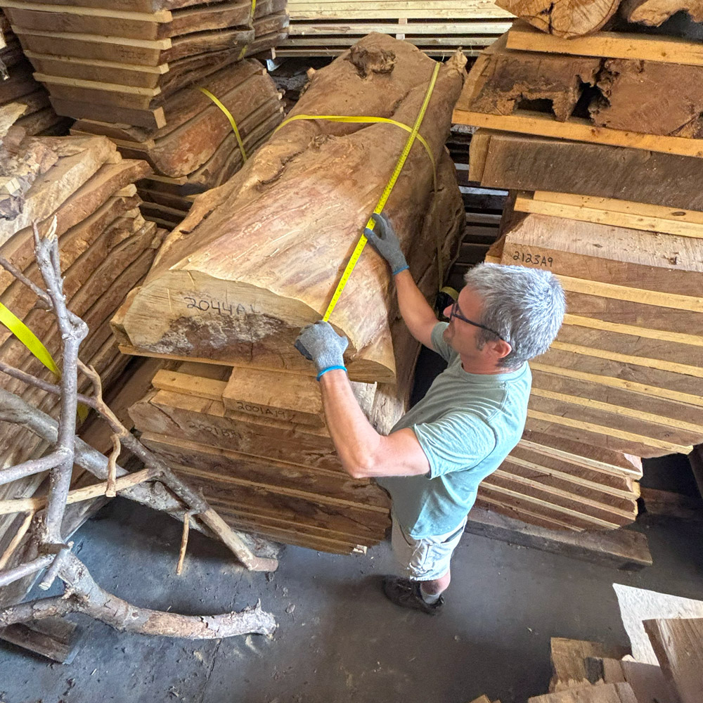 A man measures planks of raw edge wood stacked in a woodworking space.