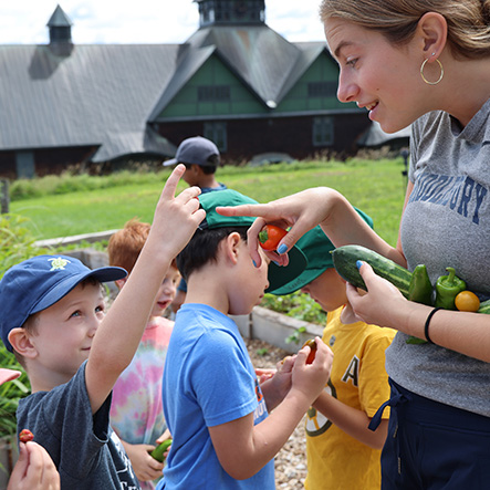 An educator helps young children examine vegetables in a garden with a large building in the background.