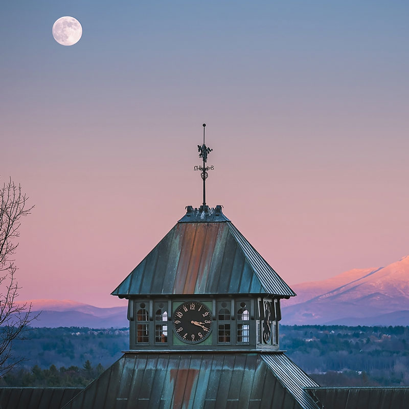 clock tower of Farm Barn in foreground. Moon rising behind it, with purply pink skies and snow capped Mount Mansfield in the background
