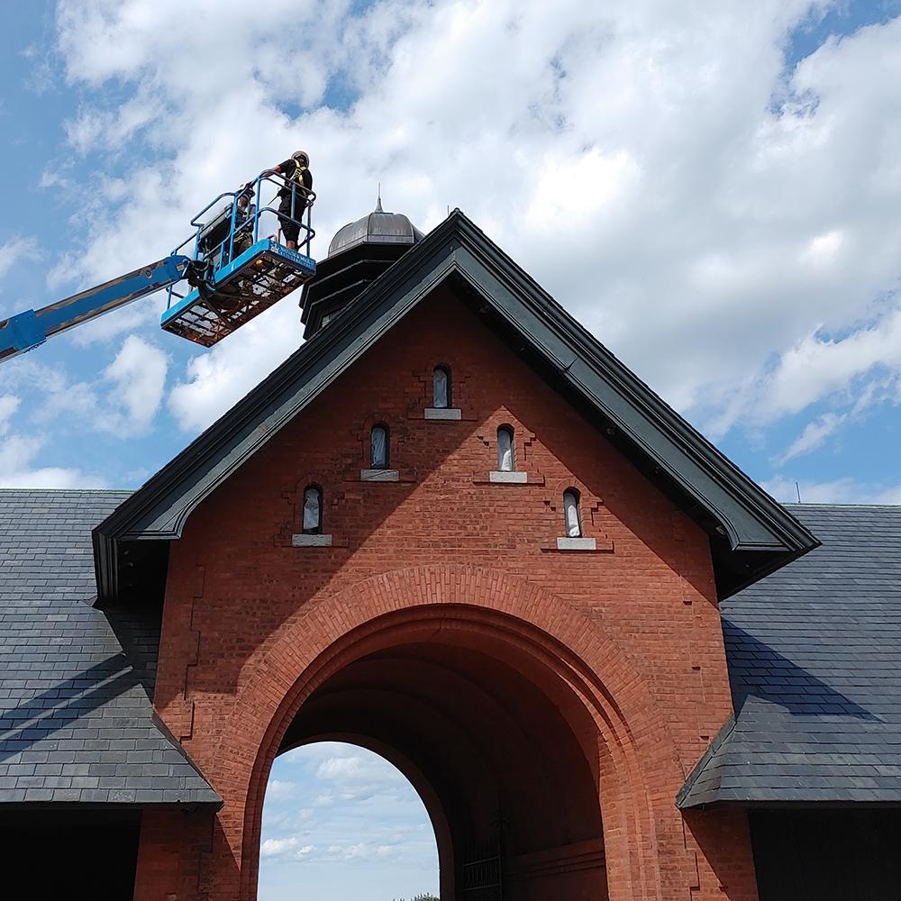 Person in a hydraulic lift performing maintenance on the roof of a red brick building with an arched entrance under a blue sky.