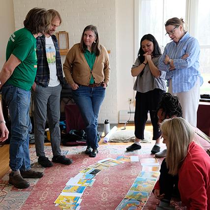 A group of adults in discussion in a living room gathered around pages of a picture book spread out on the floor
