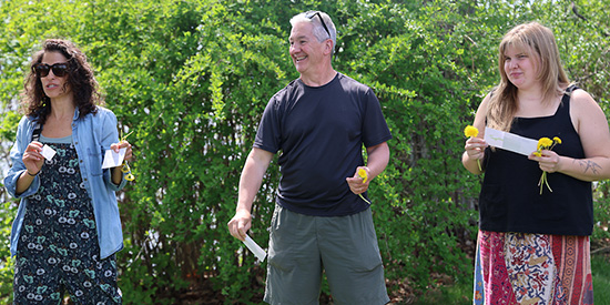 Three adults stand outdoors holding index cards and flowers during an activity