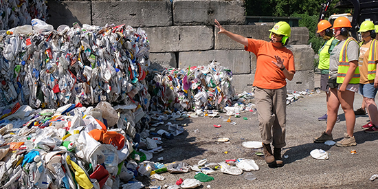 People in hard hats gesture toward compacted recycling at a facility