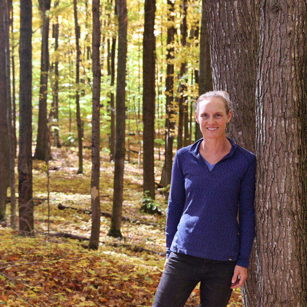 A woman in a bright yellow autumnal forest leans against a large tree