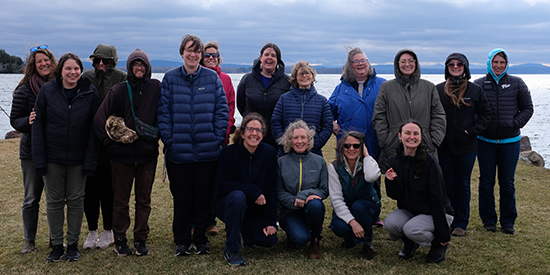 A group of 16 adults pose for a photo on a spring lakeshore