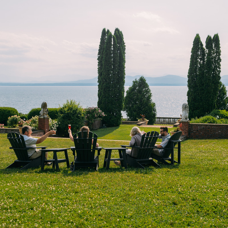 Four individuals sitting in Adirondack chairs by a scenic lake, surrounded by tall trees and a formal garden.