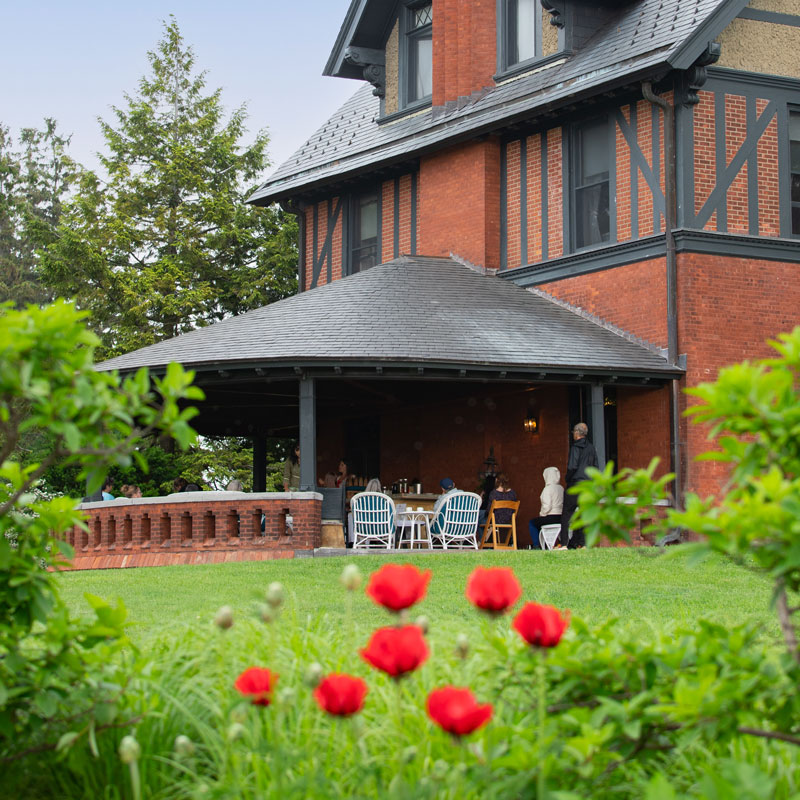 A brick building with a porch where people sit at a table. Foreground features a lush green lawn and vibrant red flowers.