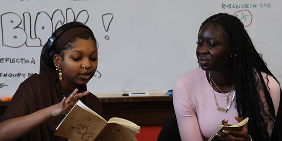 Two high school aged students sit at a table in a classroom sharing journals
