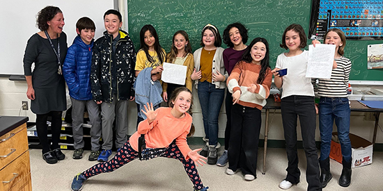 A group of young students pose with their teacher in front of a classroom chalkboard