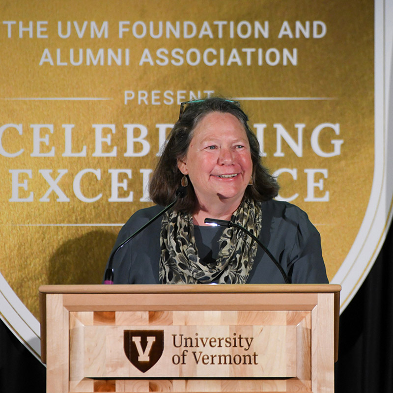 A woman with shoulder length hair stands at a UVM podium making a speech