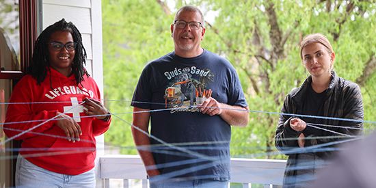 Three adults smile while holding pieces of string connected to a larger web part of a team building activity