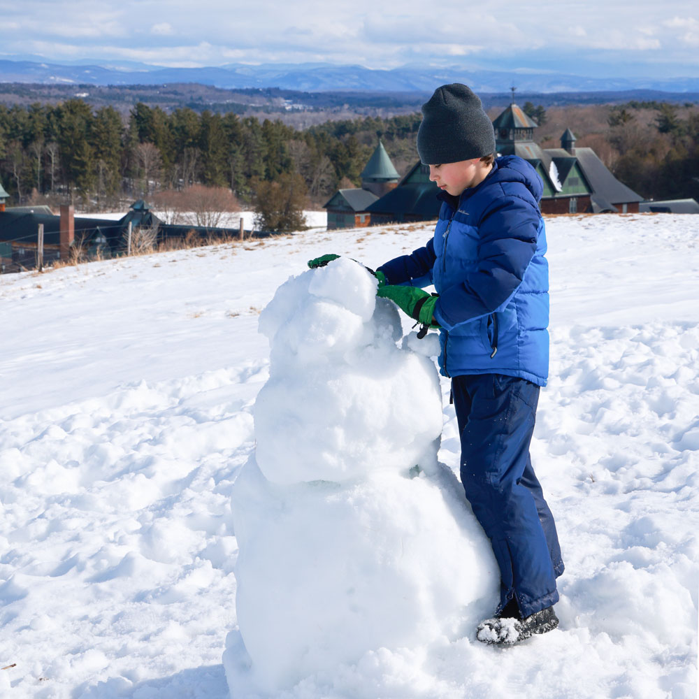 A child in winter outdoor clothes builds a snowman, the Farm Barn in the background.