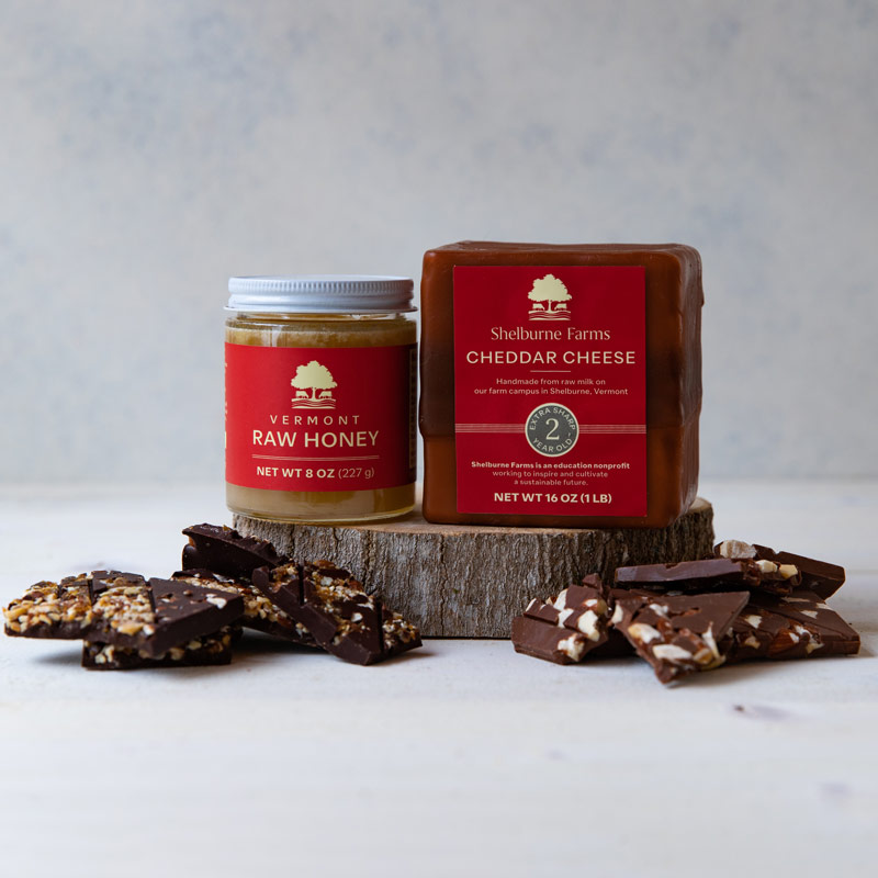 A block of Shelburne Farms Cheddar Cheese and a jar of Vermont Raw Honey are displayed on a small wooden platform. Pieces of chocolate bark with various nuts and seeds are scattered around them.