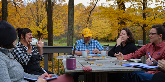 Six adults gather around an outdoor table talking with yellow foliage in background