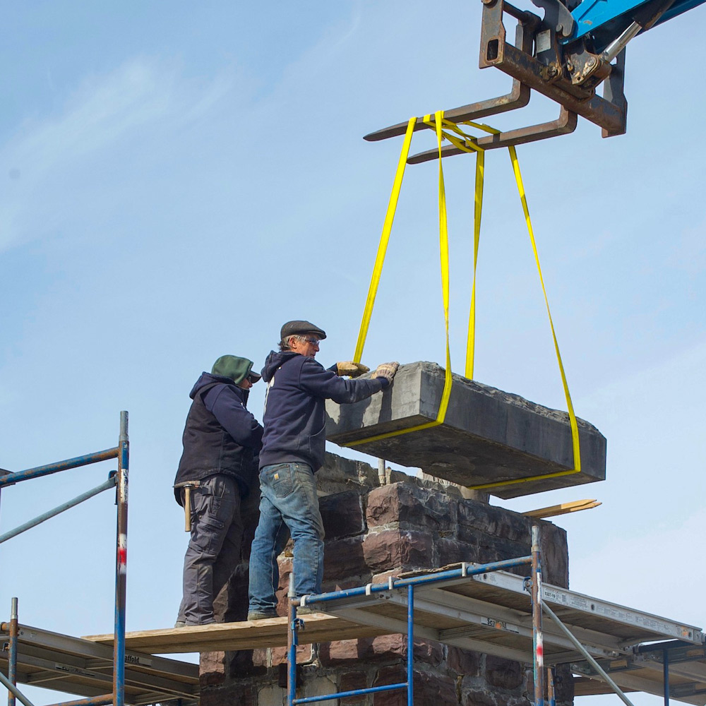 Two people are standing on scaffolding, guiding a large stone block being lowered by a crane onto a stone wall.