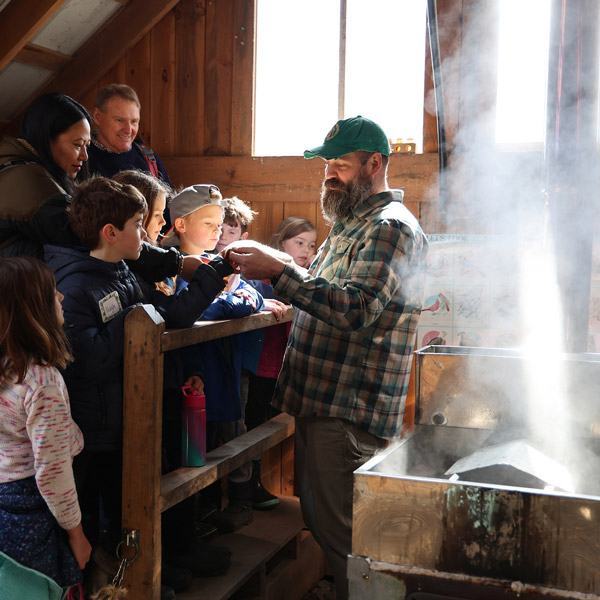 A person demonstrating the maple syrup making process to a group of attentive visitors inside a wooden sugar shack.