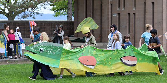 A small group of young people march across a grassy field with a large trout puppet