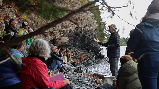 A group of people sit on a rocky lakeshore listening to a man speaking