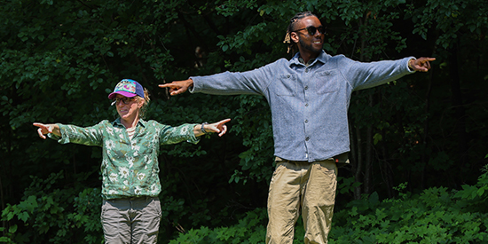 Two adults stand outdoors pointing in opposite directions and smiling during a team building exercise