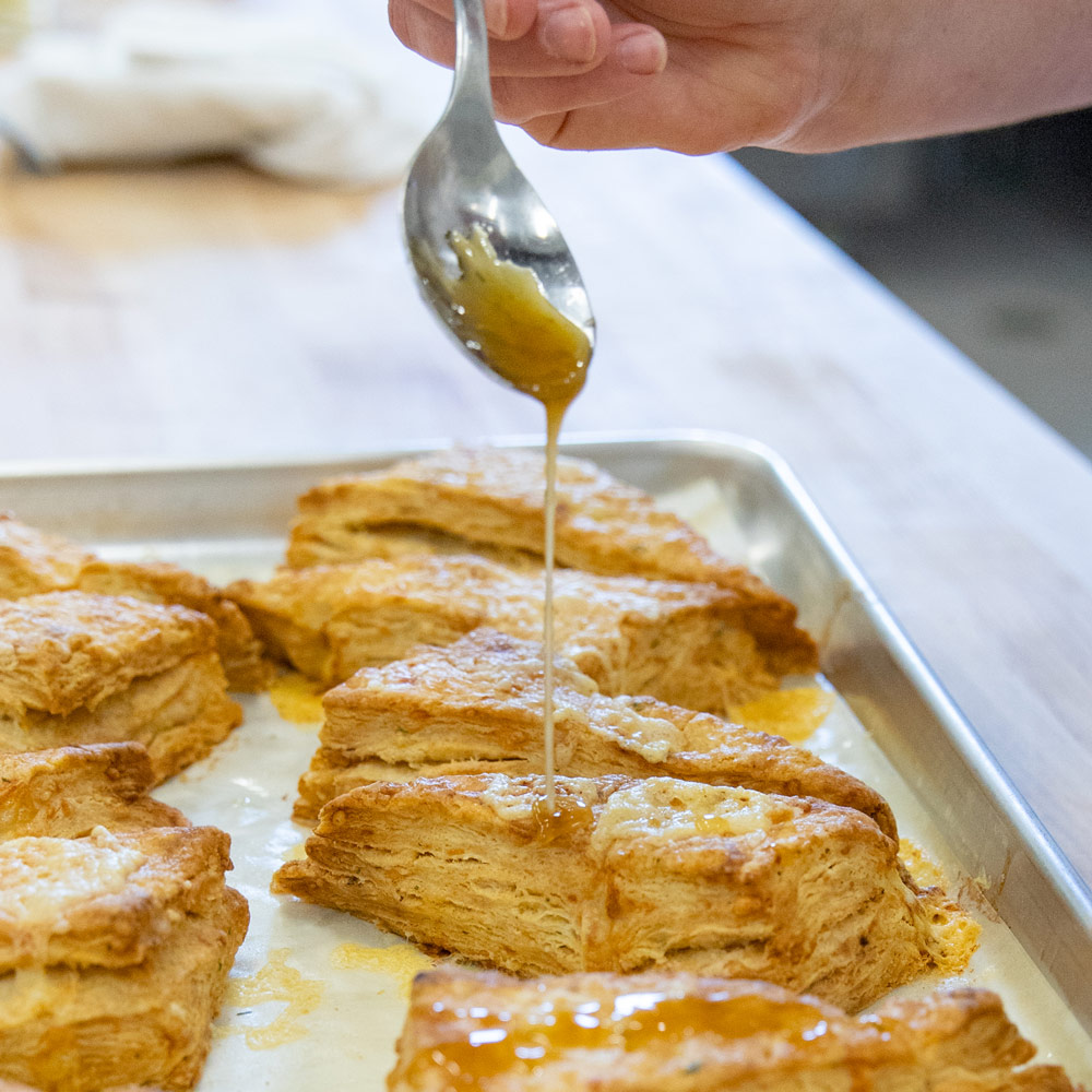 A spoon is used to drizzle maple syrup over a freshly baked tray of scones.