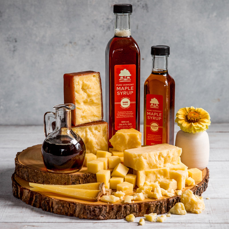 A wooden serving board with cheese and bottles of maple syrup and a vase with a flower.