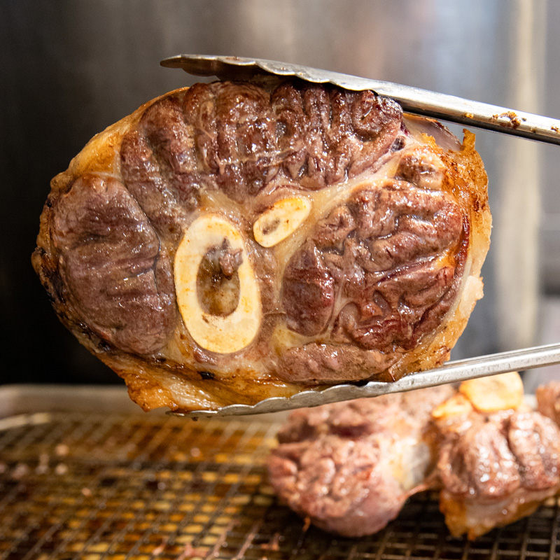 Close-up view of a roasted beef shank with a bone marrow section, glistening as it is held up by tongs.