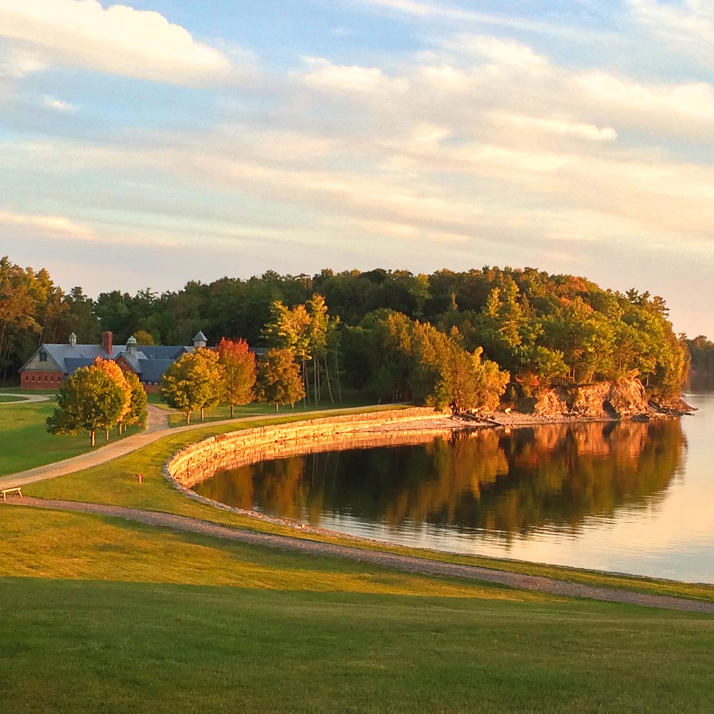 A brick building surrounded by lush trees, a curved stone shoreline, and calm water reflecting the trees and sky during sunset.