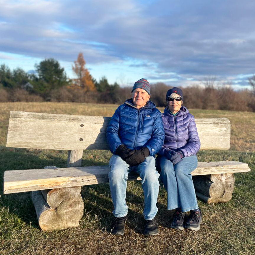 an older man and woman sit together on a rustic wooden bench surrounded by autumn grass. They are both in jeans, wearing winter coats, hats, and mittens.
