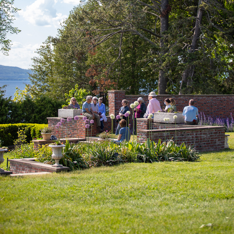 A group of adults gather by a low brick wall in a lakeside garden.