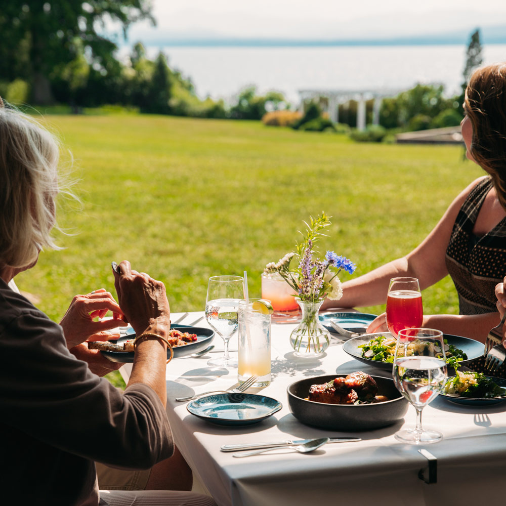 Two people dining at a table outdoors with scenic views of a grassy lawn, gardens, and lake.