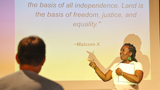 A woman speaks in front of a projector screen displaying a Malcom X quote