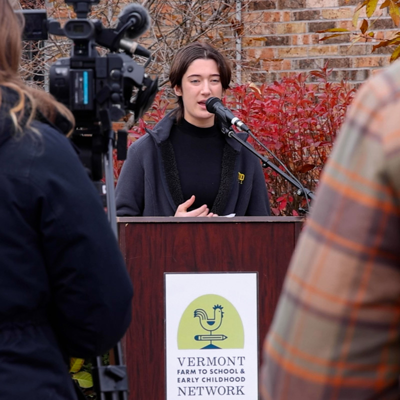 A student speaks at a podium with a news camera and audience in the foreground